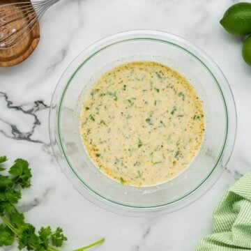 A smooth and creamy yellow marinade with green seasonings sits in a glass bowl.