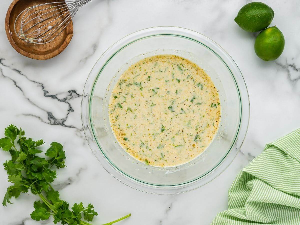 A smooth and creamy yellow marinade with green seasonings sits in a glass bowl.