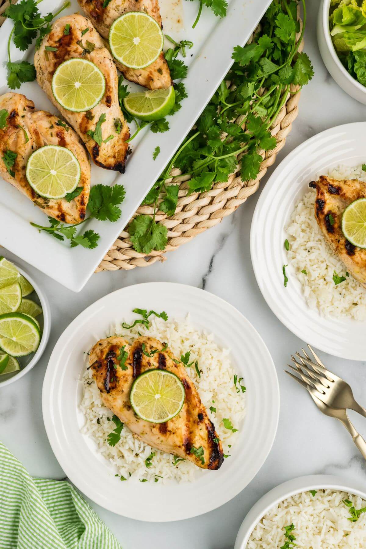 A long platter and white plates have multiple chicken breasts topped with lime and cilantro.