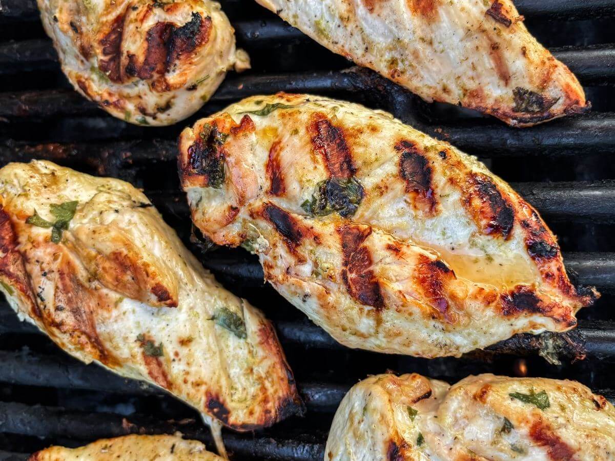 Six chicken breasts with dark grill marks and green garnish sit on a grill.