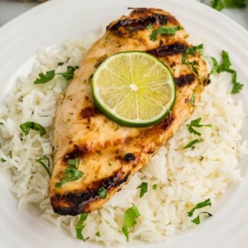 A plate full of white rice with cilantro is topped with a grilled chicken breast with lime.