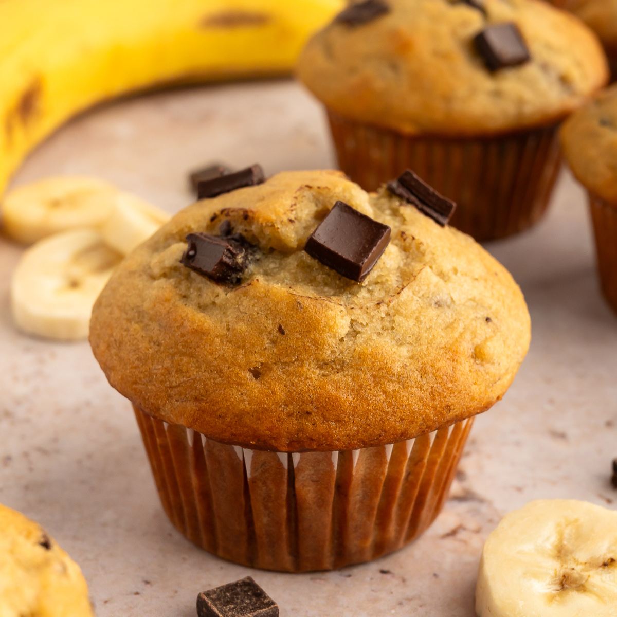 Banana chocolate chunk muffin surrounded by chocolate chunks, banana slices and a ripe banana.