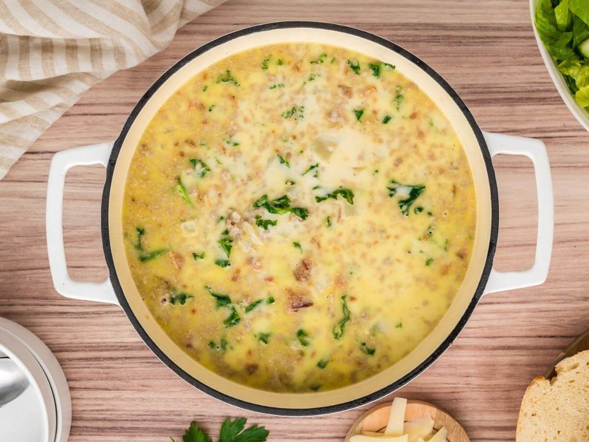 Creamy potato based soup shows pieces of green kale, bacon and sausage in a white Dutch oven pot.