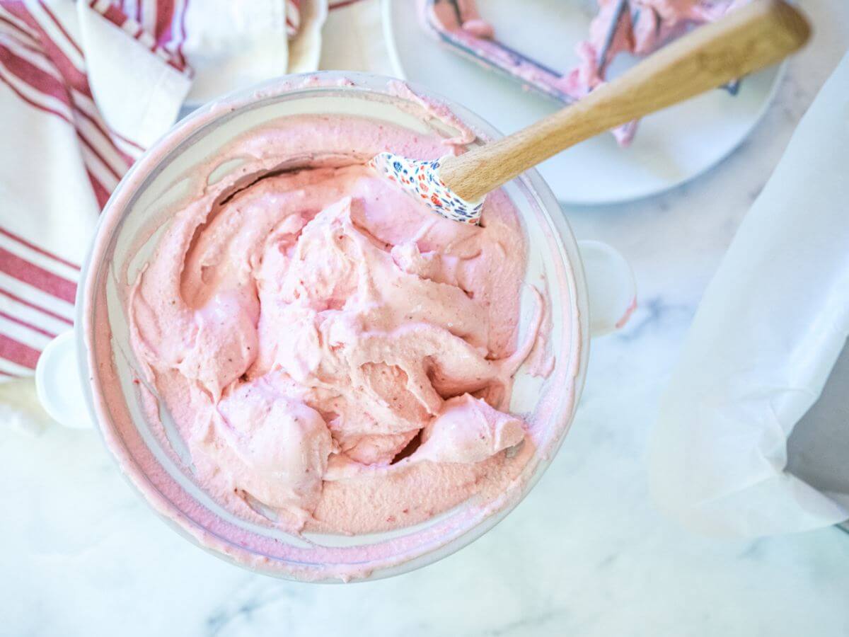 Thick and creamy light pink strawberry ice cream in ice cream maker with spatula. 