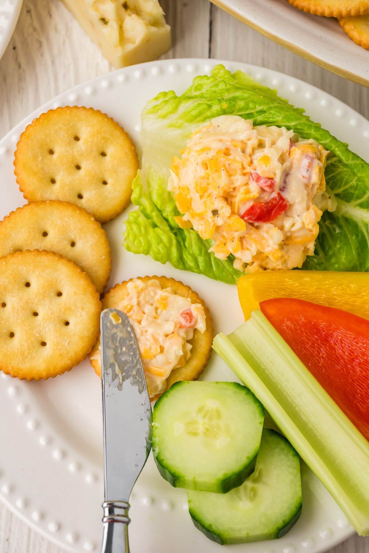 Spreading the pimento cheese onto a cracker on a plate with some veggies on the side.