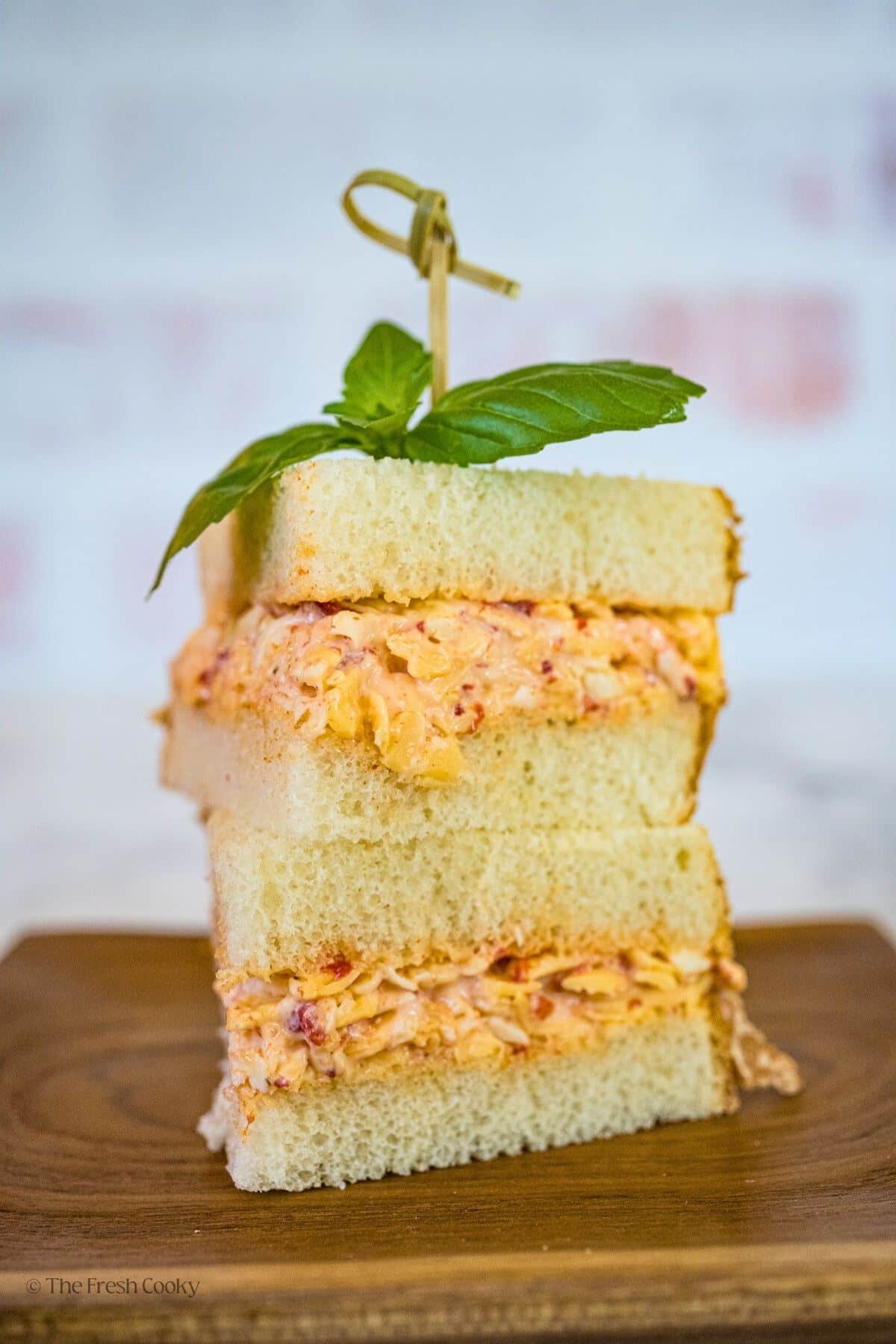 Pimento cheese sandwich slices on a wooden plate with a sandwich pick in the top and a sprig of basil. 