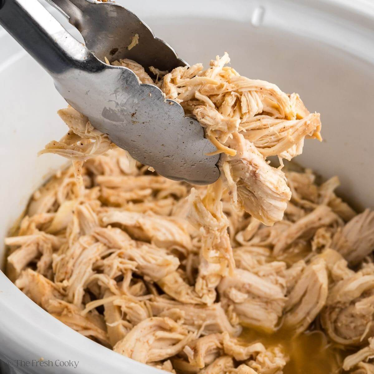 Tongs picking up some shredded chicken from a slow cooker full of chicken.
