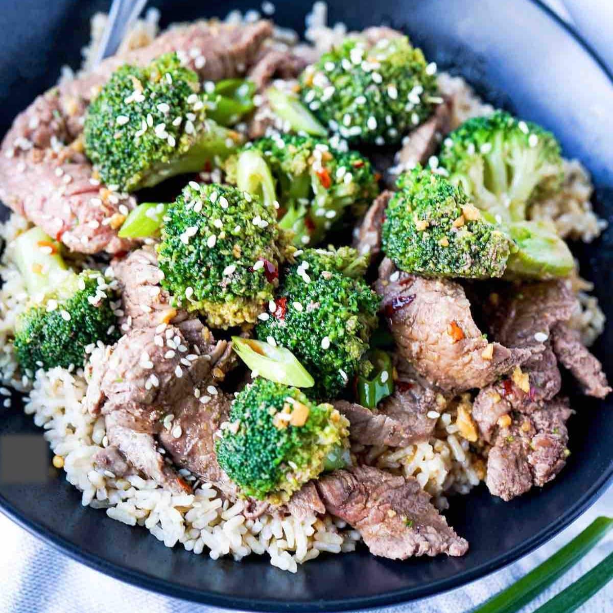 Beef and Broccoli in a black bowl with rice and sauce on top.