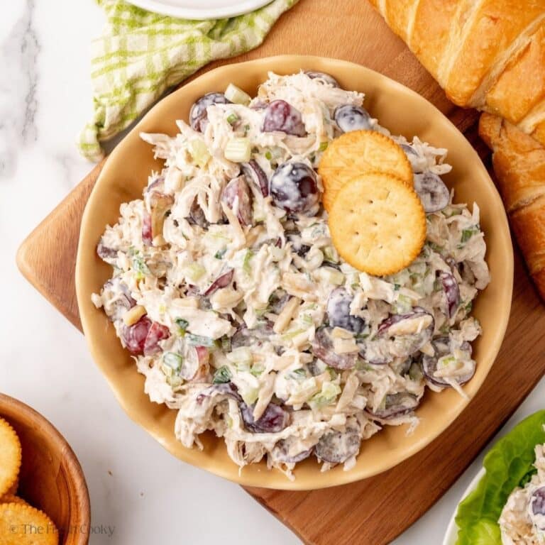 Chicken salad with grapes in a pretty bowl surrounded by croissants, crackers and lettuce.