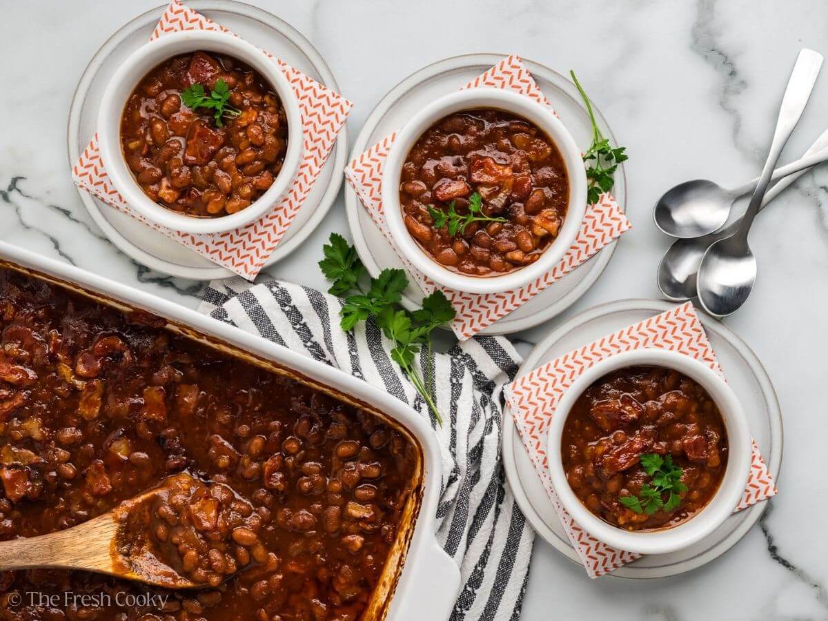 3 servings of cowboy baked beans in small bowls next to the large serving dish full of beans. 