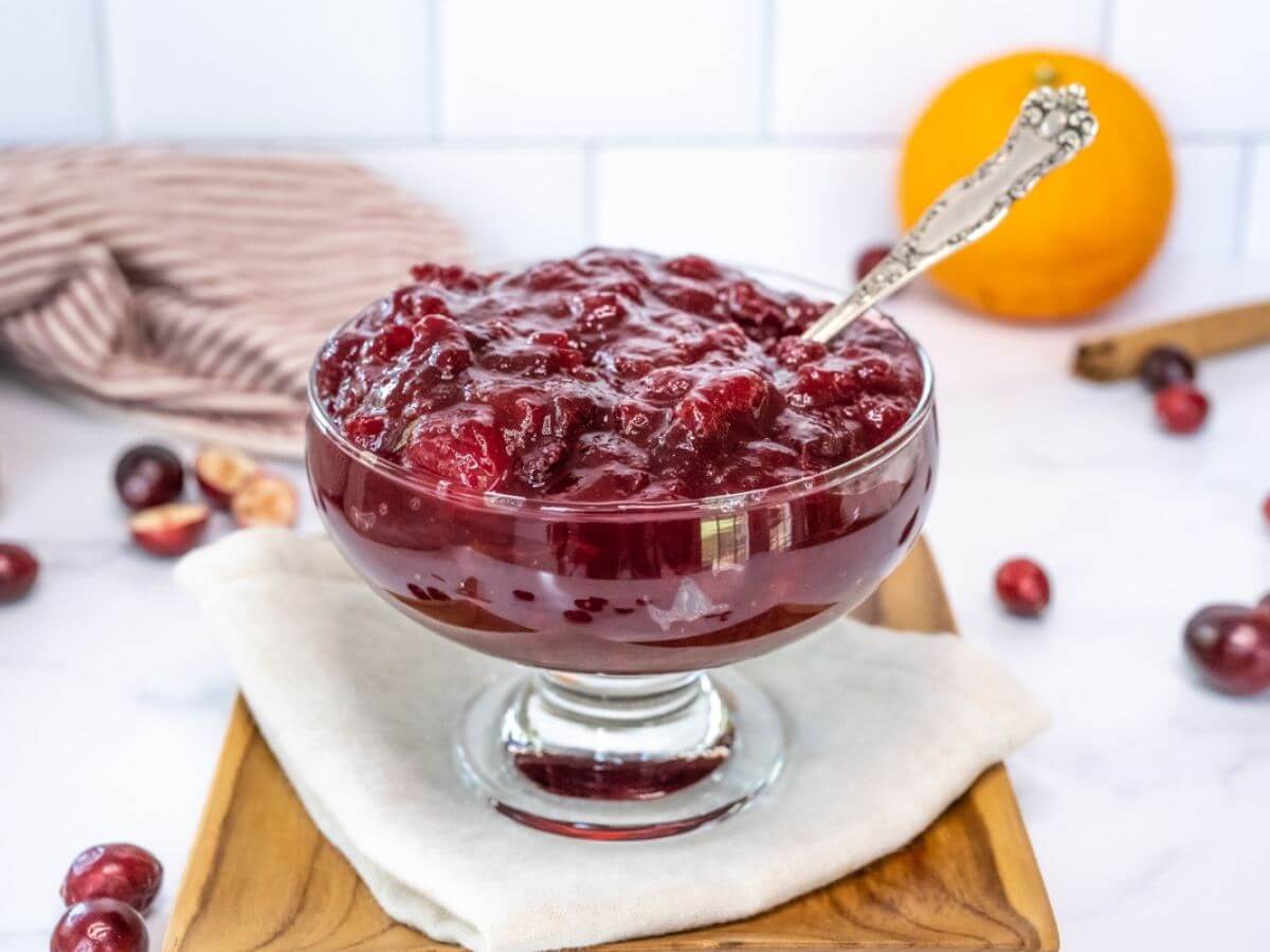 Glass serving dish of cranberry sauce on a wooden board with fresh cranberries and an orange in the background.