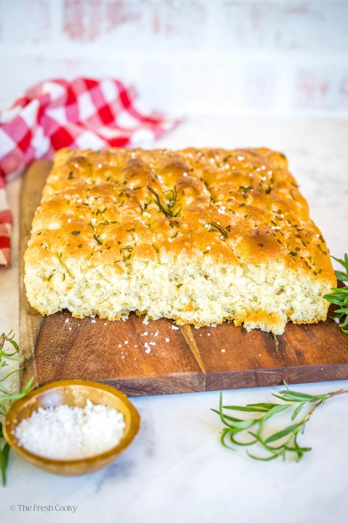 Focaccia bread on a wooden cutting board with salt and rosemary sprigs around it.