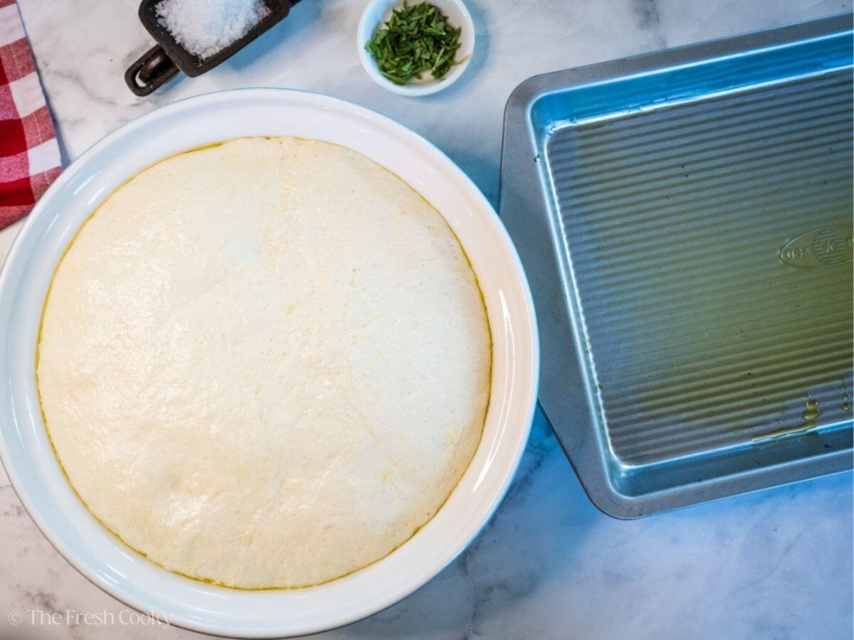 Risen focaccia dough in a white bowl next to a baking dish lined with olive oil.