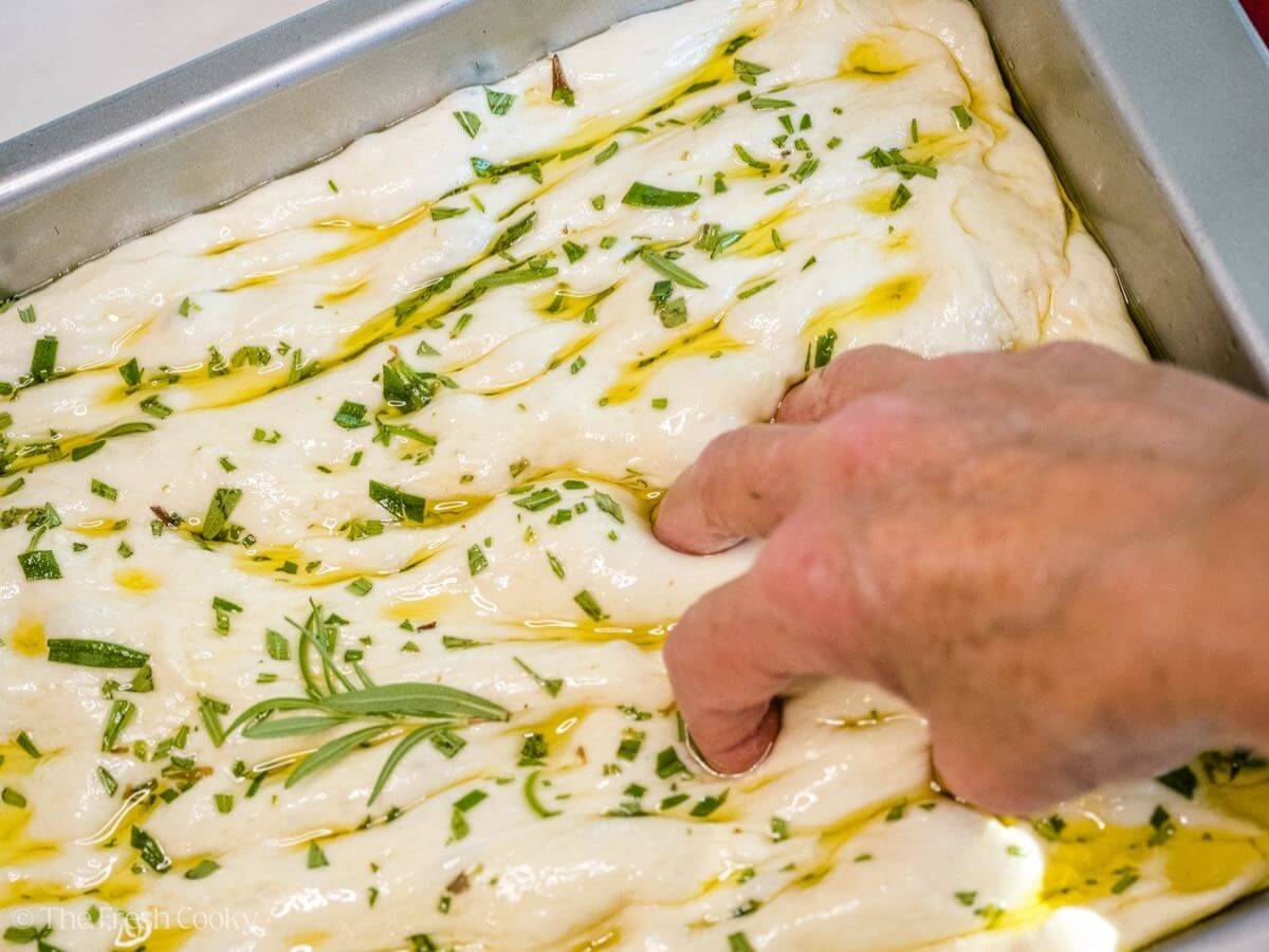 Fingertips pressing into the focaccia dough to create a dimple pattern.