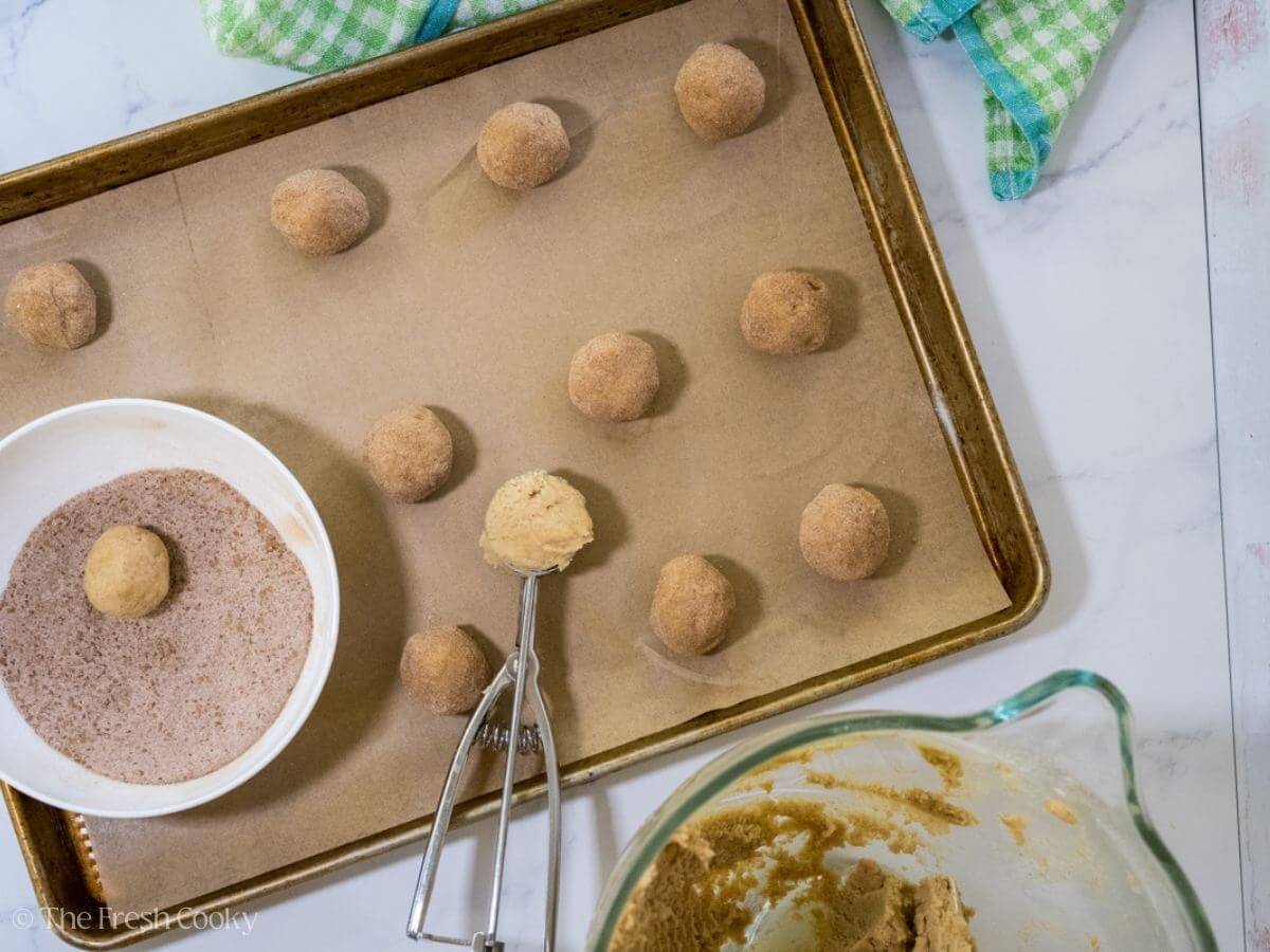 Overhead shot of snickerdoodle cookie dough balls on a sheet pan while some are being rolled in sugar.