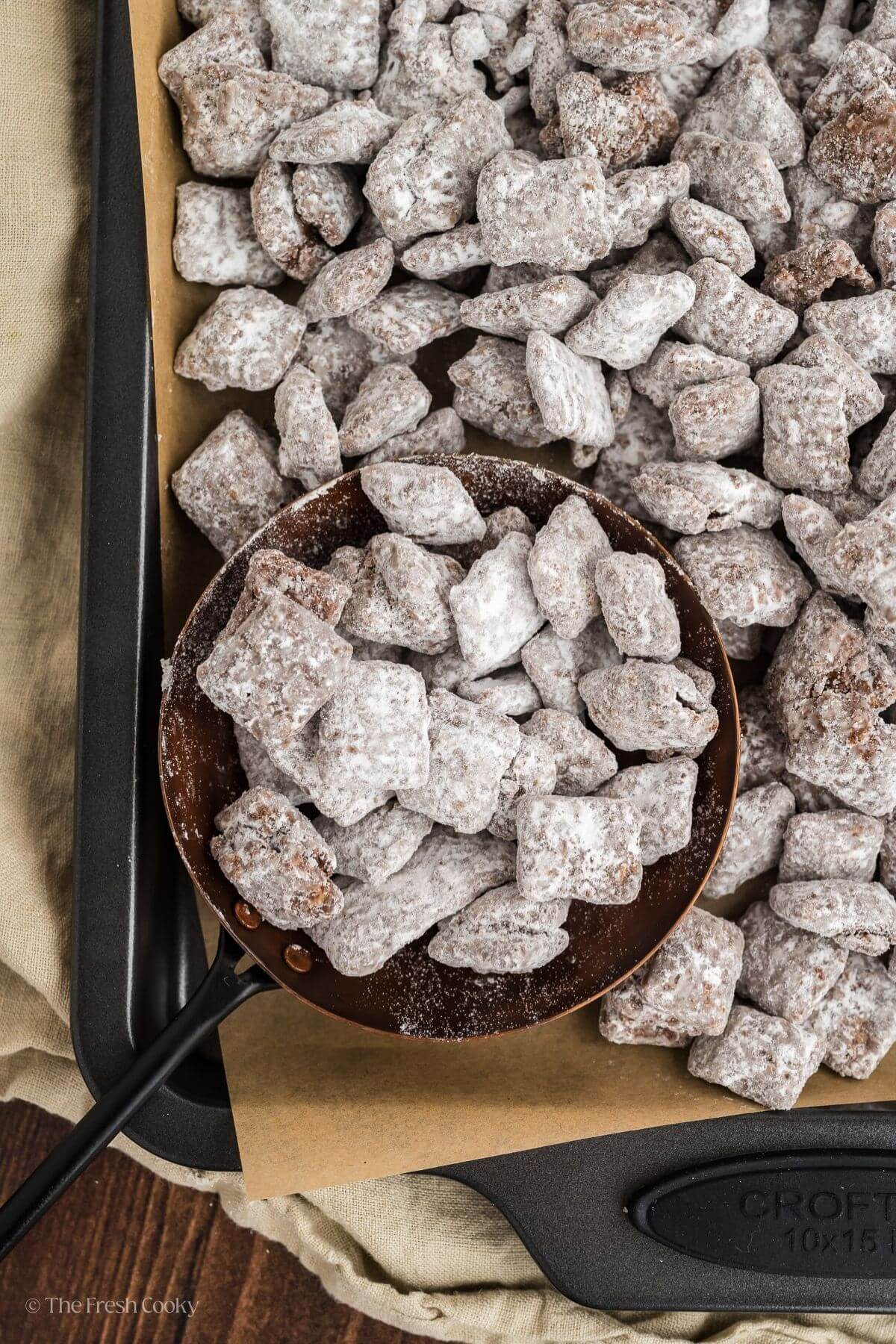 Puppy chow on a baking sheet with a measuring cup scooping some up.