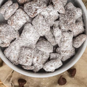 Puppy chow served in a white bowl on a cloth with chocolate chips on it.