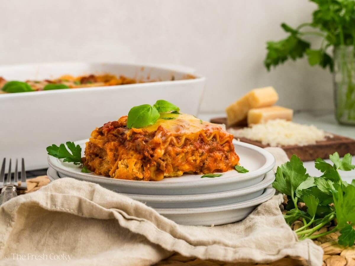A slice of cheesy lasagna garnished with basil on a plate, with a baking dish and cheese in the background surrounded by herbs.