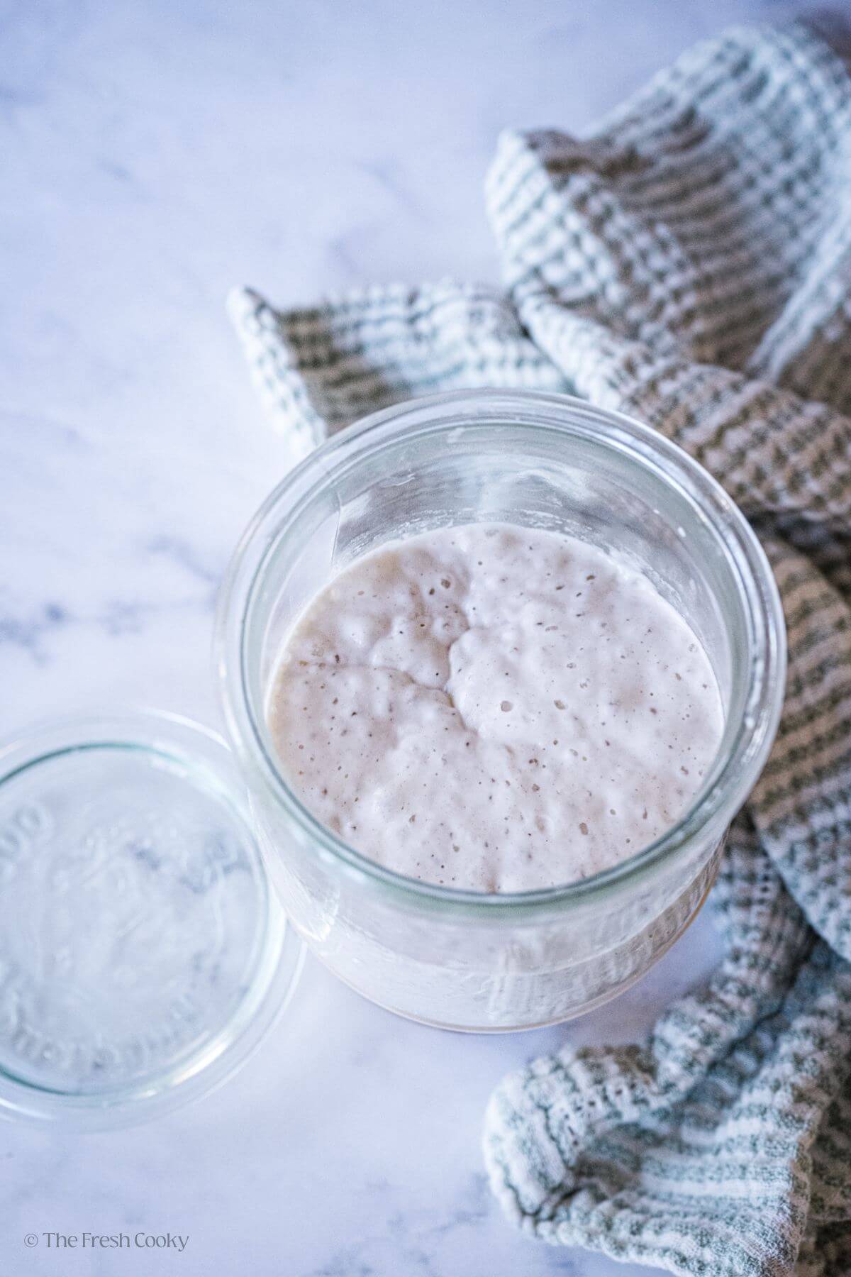 Bubbly sourdough starter in a glass jar next to a kitchen towel.