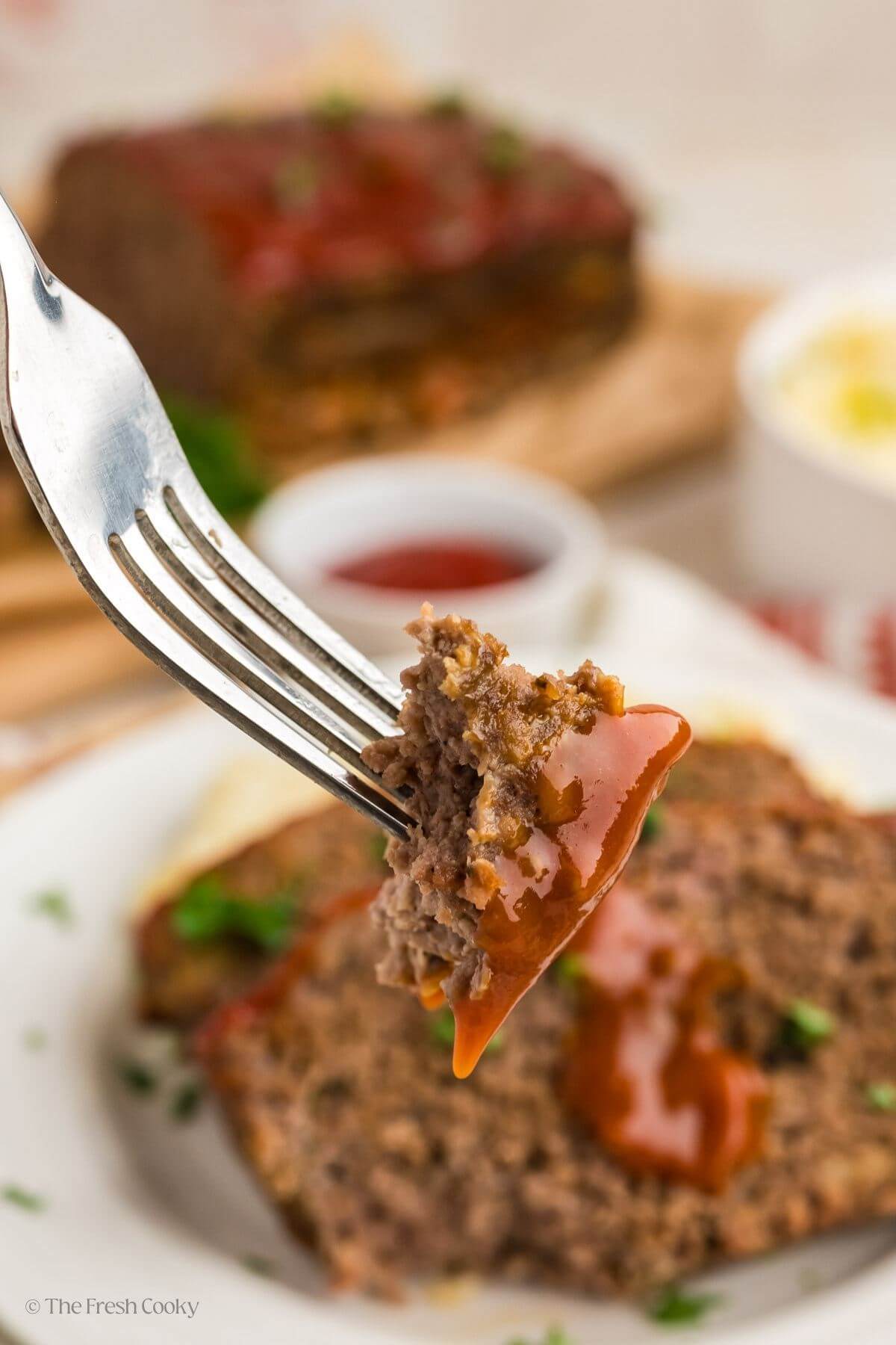 Fork with a piece of meatloaf on it with the plate of more meatloaf in the background.