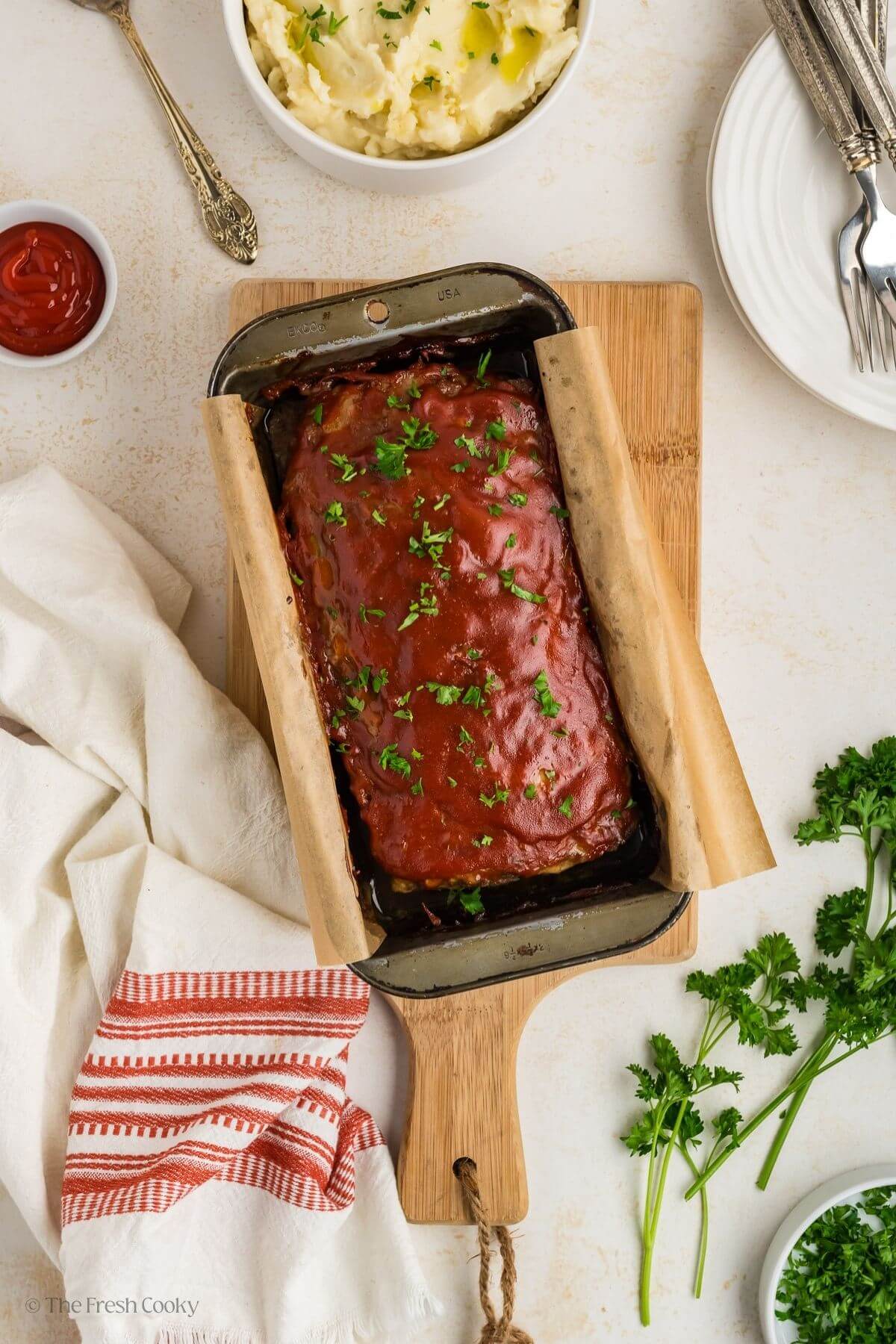 Overhead shot of baked meatloaf in a loaf pan on a wooden board with mashed potatoes on the side.