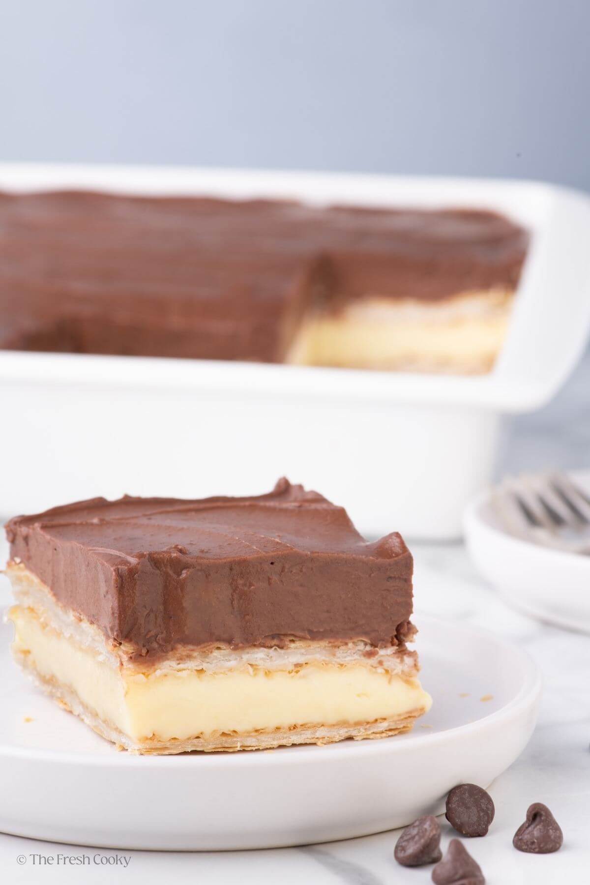Slice of chocolate eclair cake on a white plate with whole cake in the background.