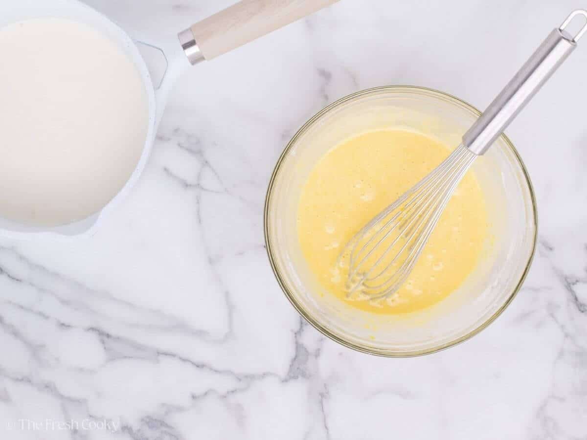 Egg yolk mixture in a glass mixing bowl next to the warm milk mixture in a saucepan.