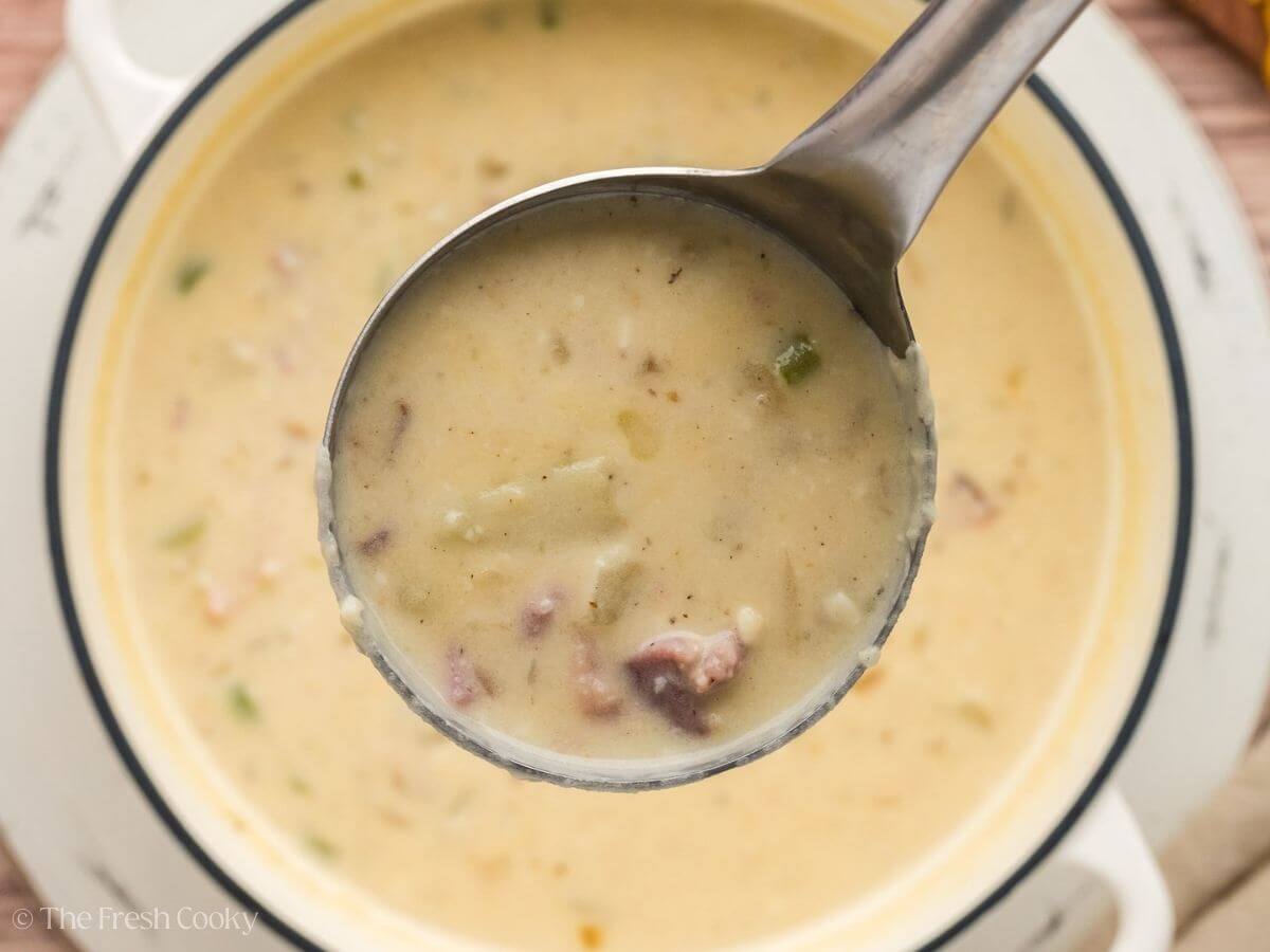 Metal ladle holding a scoop of ham and potato soup up to the camera with the pot of soup below it.
