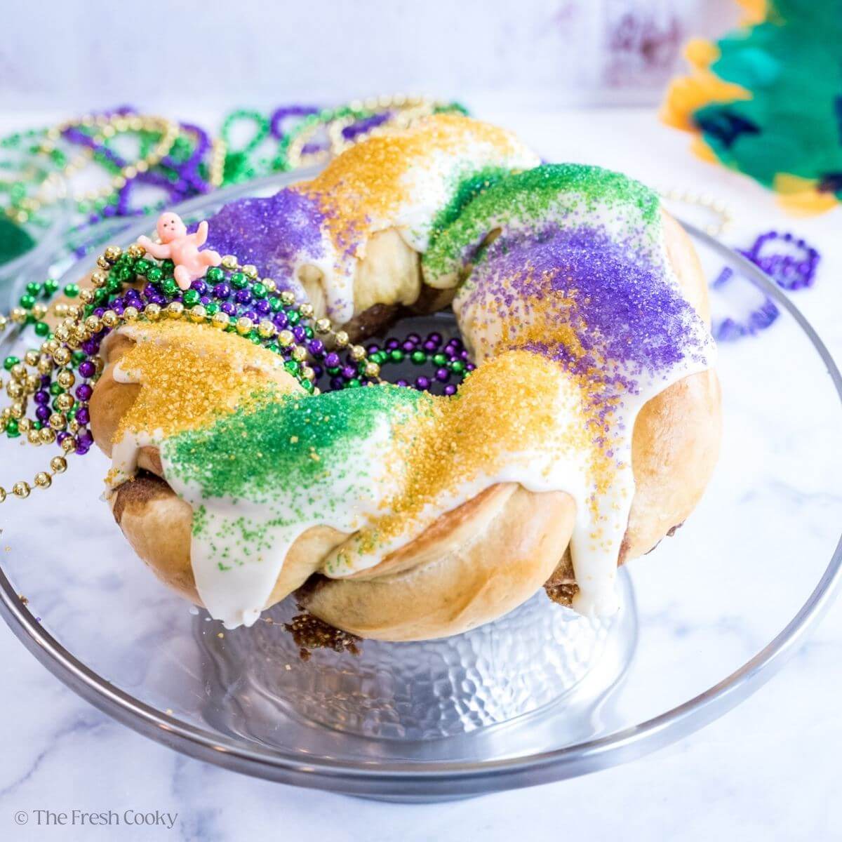 Traditional king cake on a cake stand with mardi gras necklaces draped over it and a plastic baby on top.