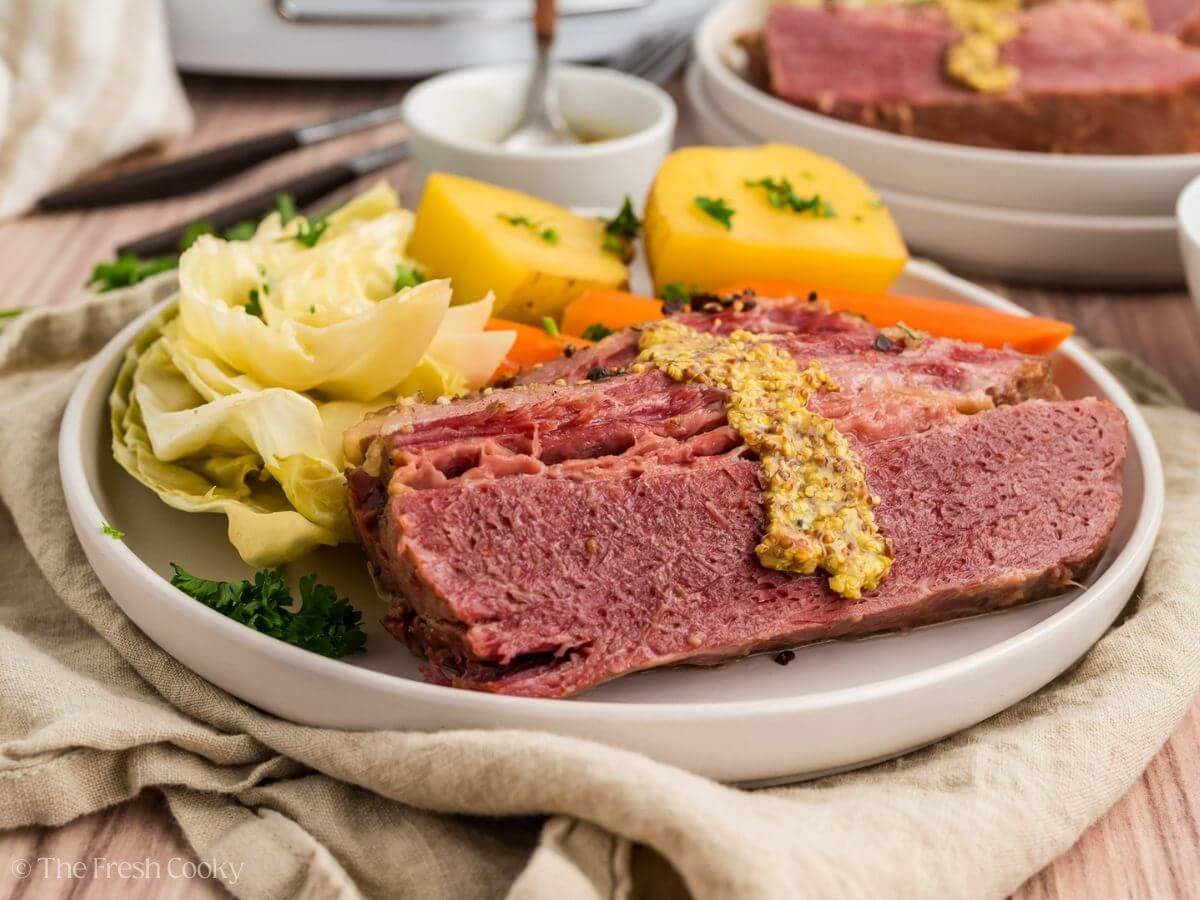 A plate of sliced corned beef topped with whole grain mustard, accompanied by boiled cabbage, potatoes, and carrots. The meal is garnished with parsley and served with a small bowl of mustard on the side.