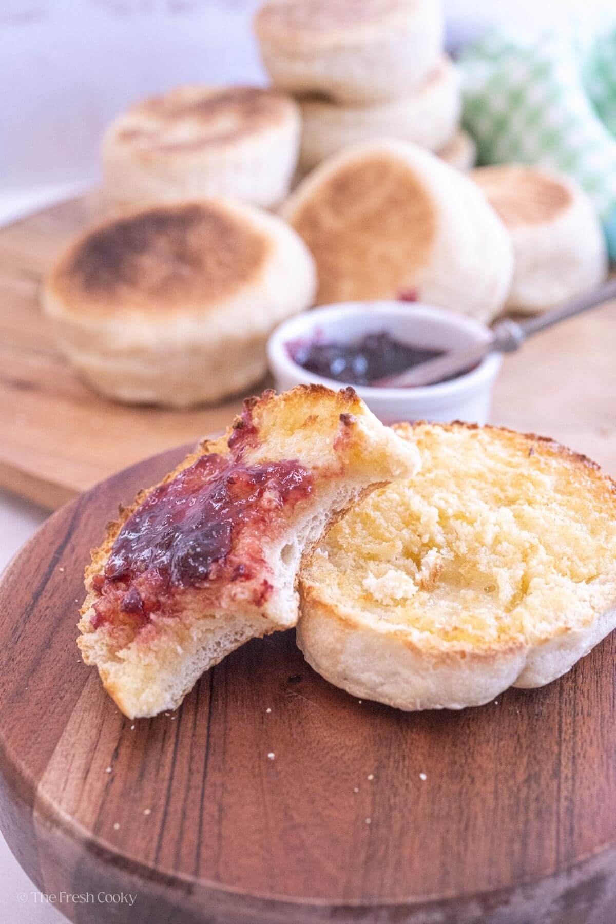 Sliced sourdough English muffin on a wooden board with jam on one half and two bites missing.