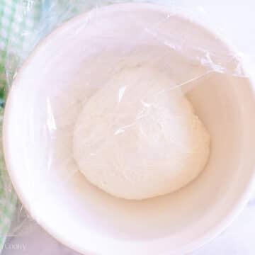 Covering the English muffin dough with plastic wrap in a mixing bowl.