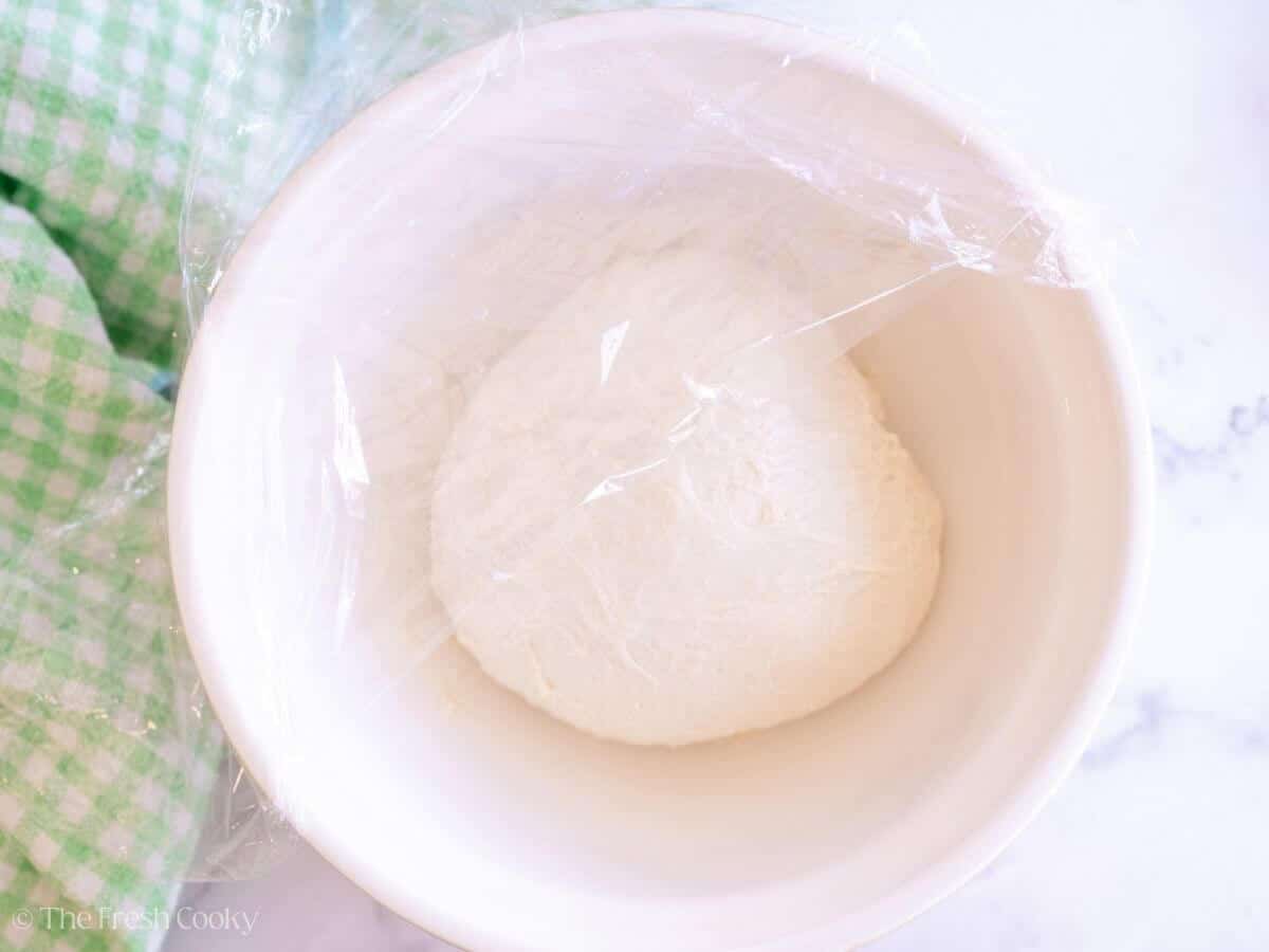 Covering the English muffin dough with plastic wrap in a mixing bowl.