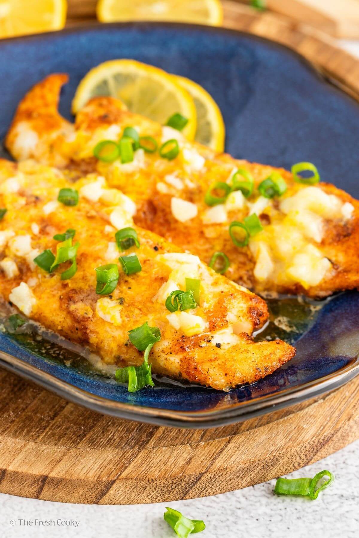Two pieces of air fried tilapia on a plate with lemon butter sauce and green onions on top.