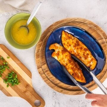 Using tongs to plate the air fryer tilapia on a plate with lemon butter and green onions next to it.