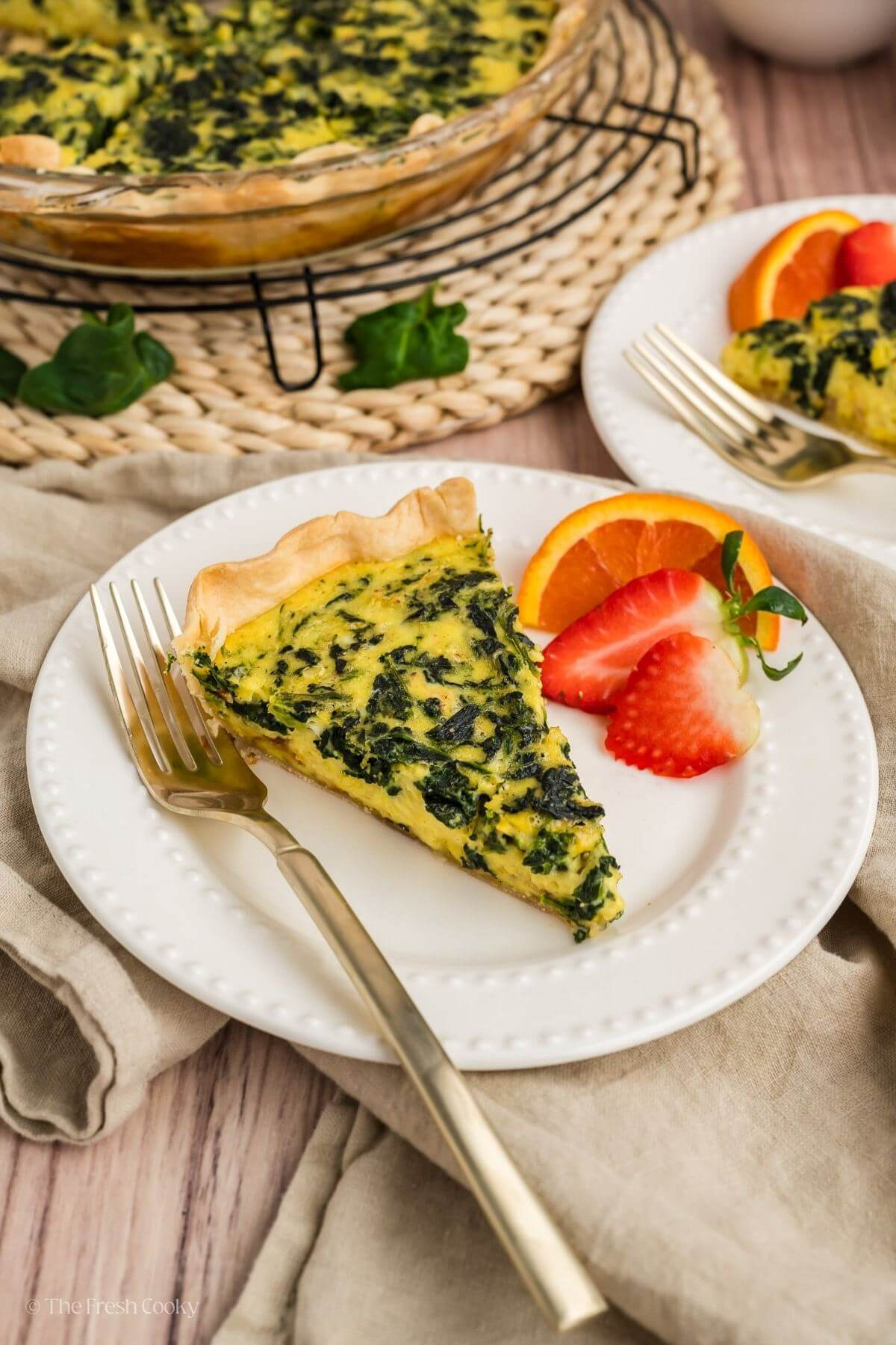 Slice of quiche florentine on a white plate with fresh fruit on the side.