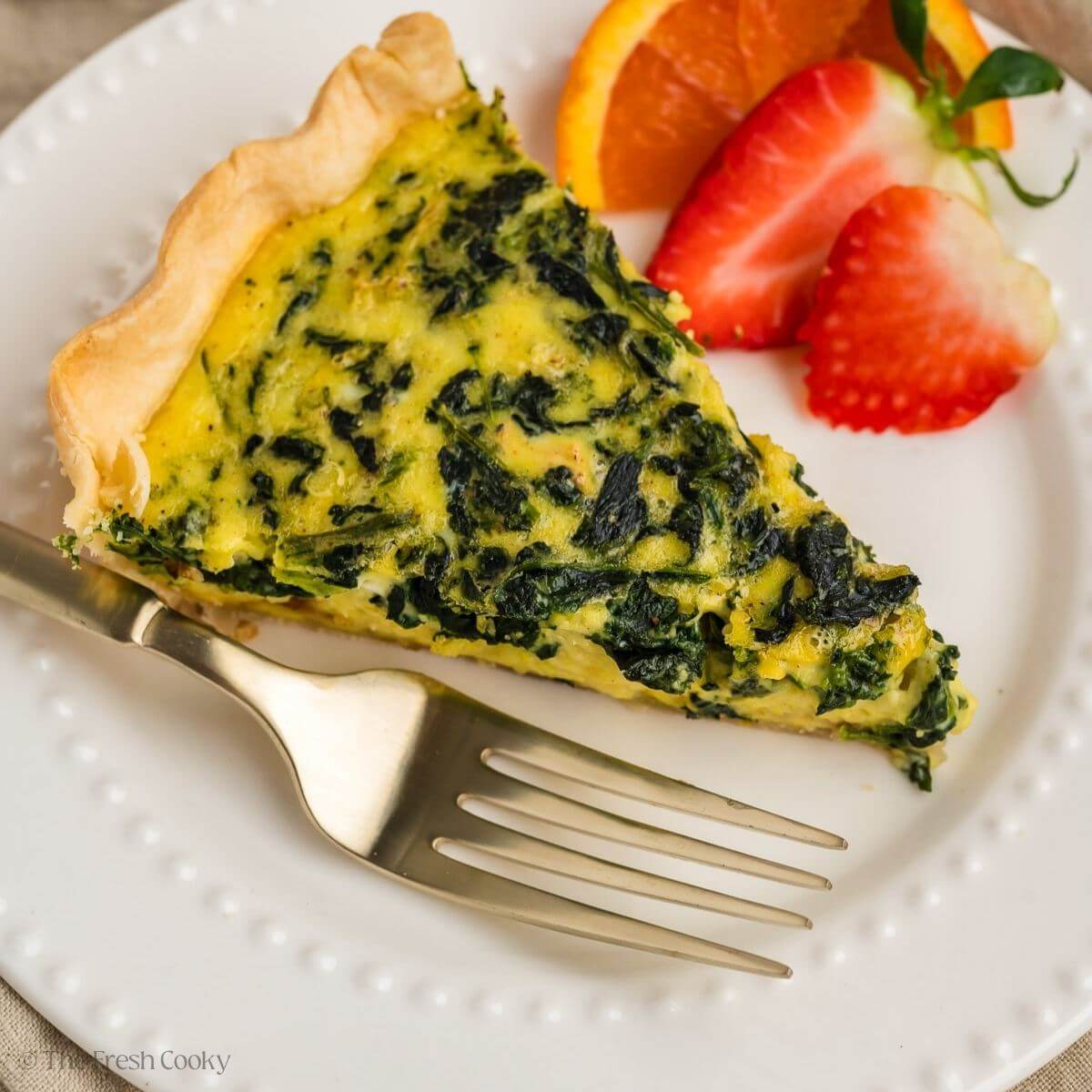 Close up shot of a slice of spinach quiche on a white plate with a side of fruit.