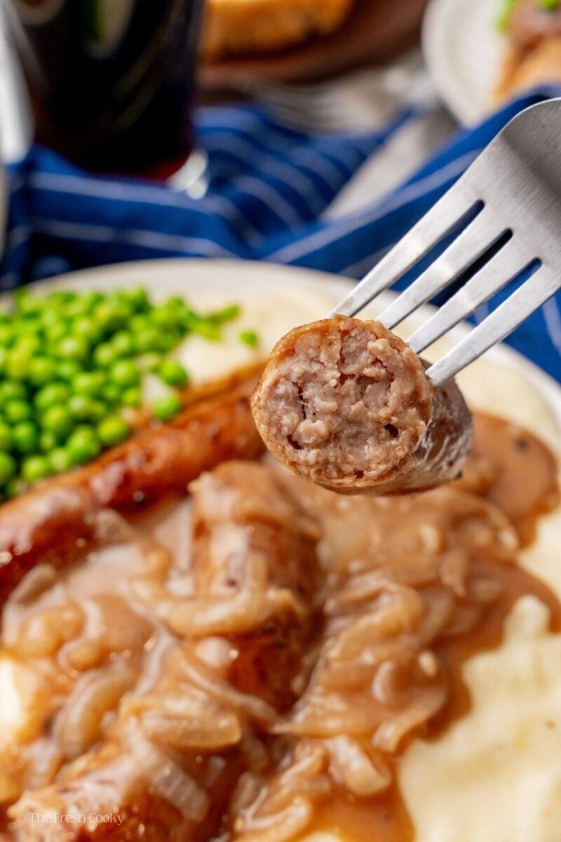 A fork taking a bite of sausage with the bangers & mash in the background on a plate with peas.