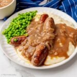 Sausage "bangers" and mash (mashed potatoes) on a plate with onion gravy and fresh peas.