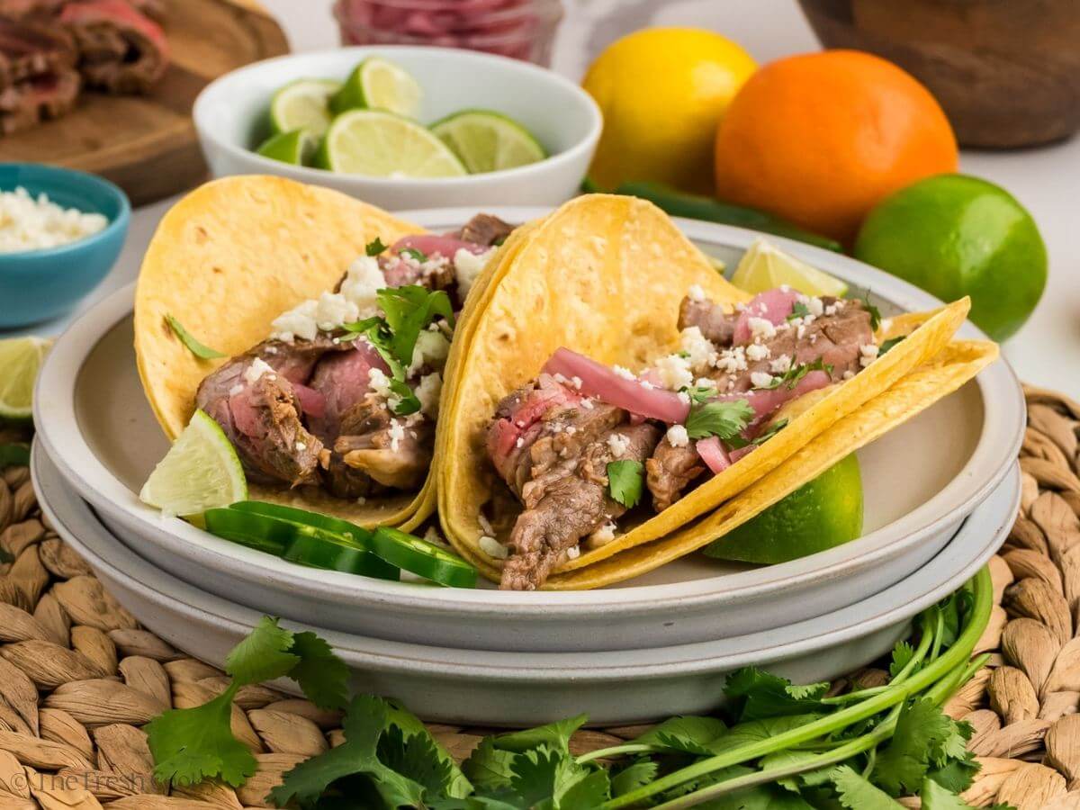 Two carne asada tacos on a plate topped with cotija cheese and pickled onions.