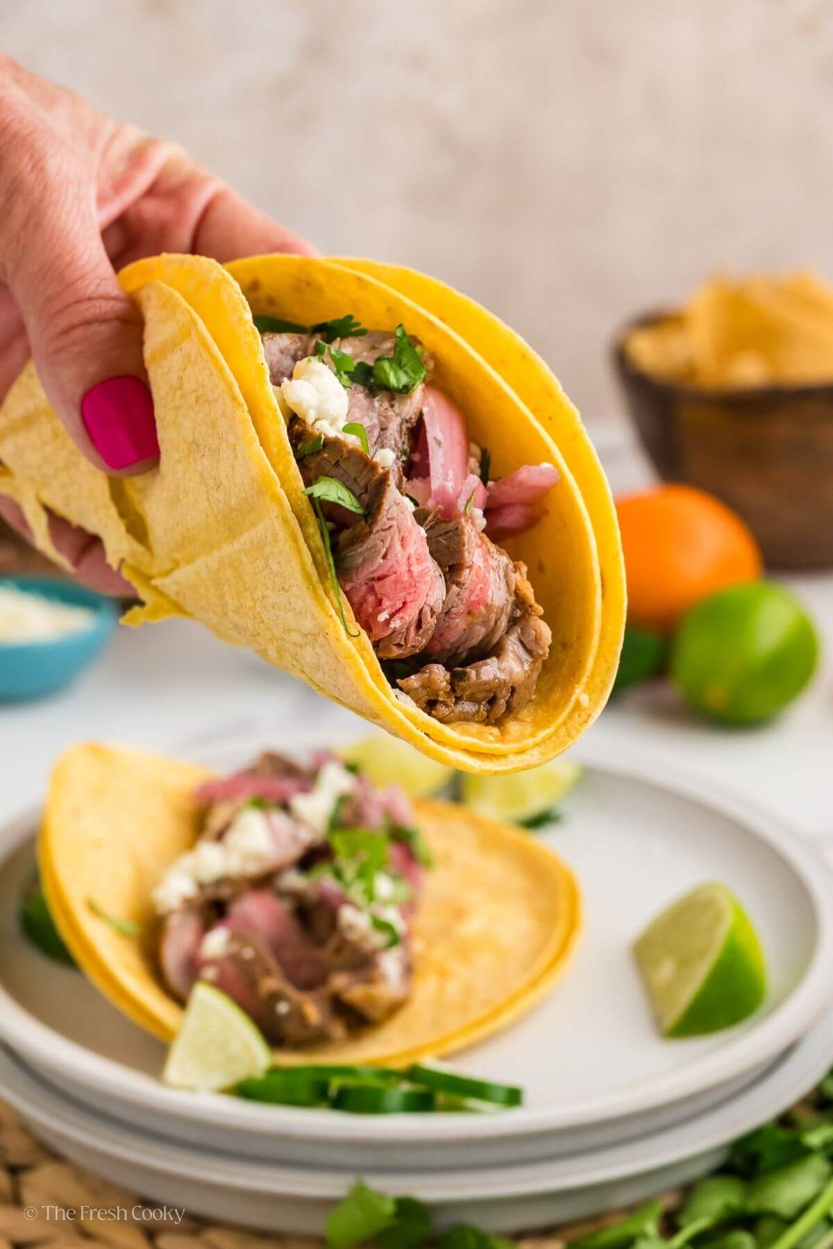 Hand holding a carne asada taco with another on a plate in the background.
