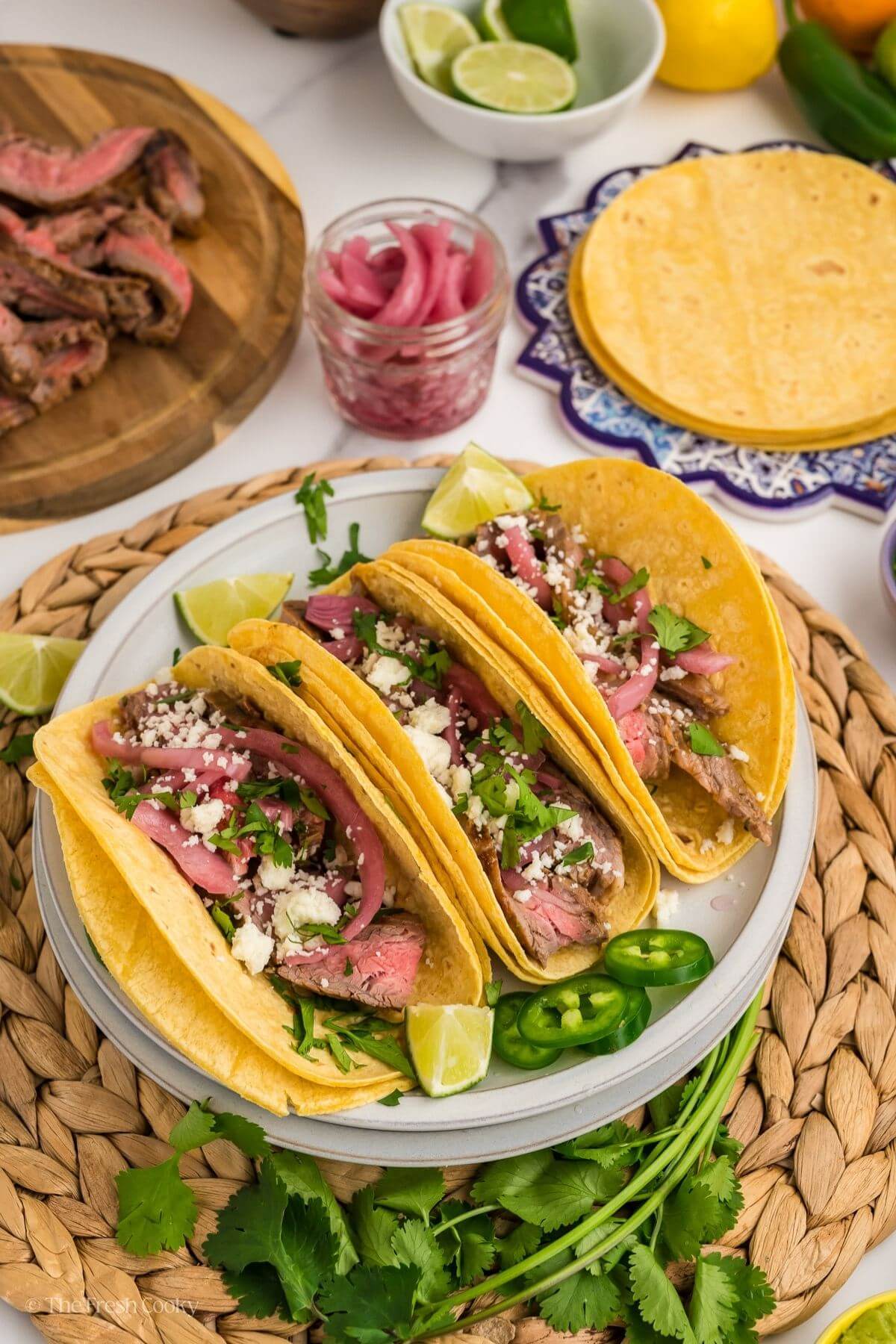 Three carne asada tacos on a plate with lime wedges and jalapeño slices around.