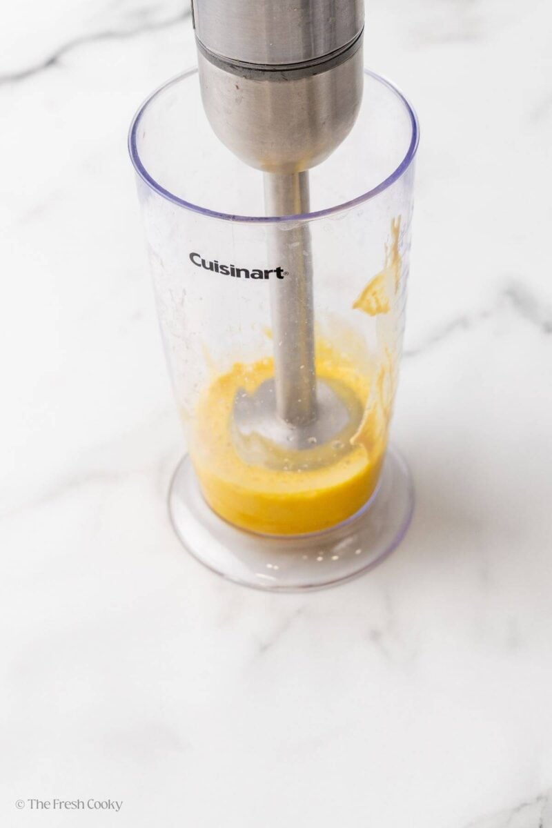 Immersion blender blending egg yolks in a plastic container.