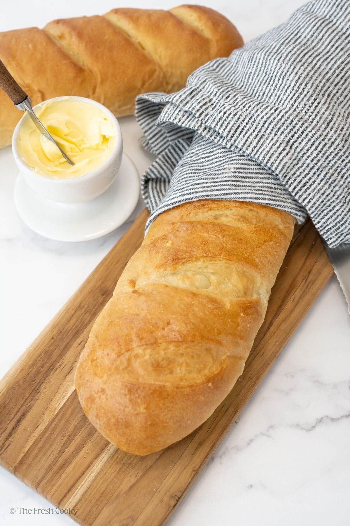 Two French bread loaves with one on a wooden board covered slightly with a linen.