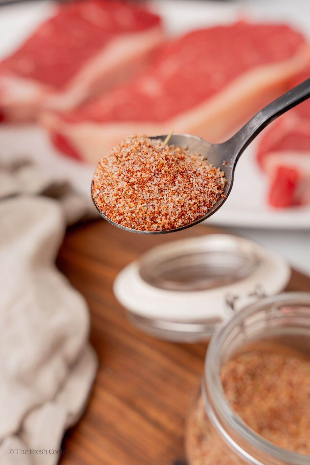 Spoon with steak seasoning on it and raw steaks in the background.