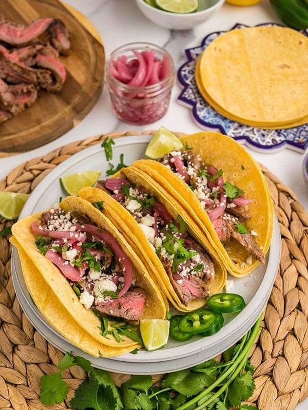 Three carne asada tacos on a plate with lime wedges and jalapeño slices around.