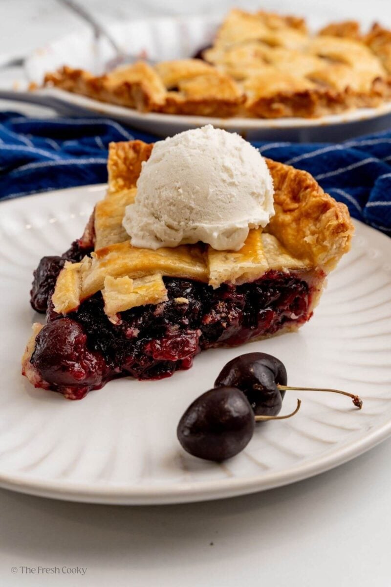 Slice of cherry pie on a white plate with ice cream on top.