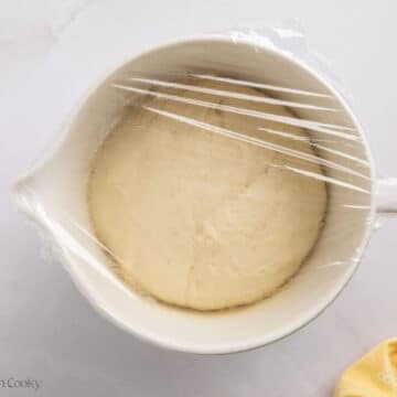 Bread dough in a white mixing bowl covered with plastic wrap to rise.