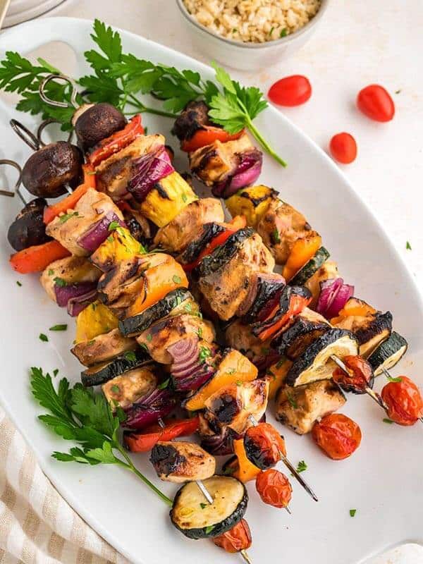 Five full shish kabobs with chicken and colorful vegetables laying diagonally next to a bowl of rice.