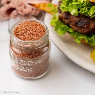 Small jar of burger seasoning next to a plate with a burger on it.